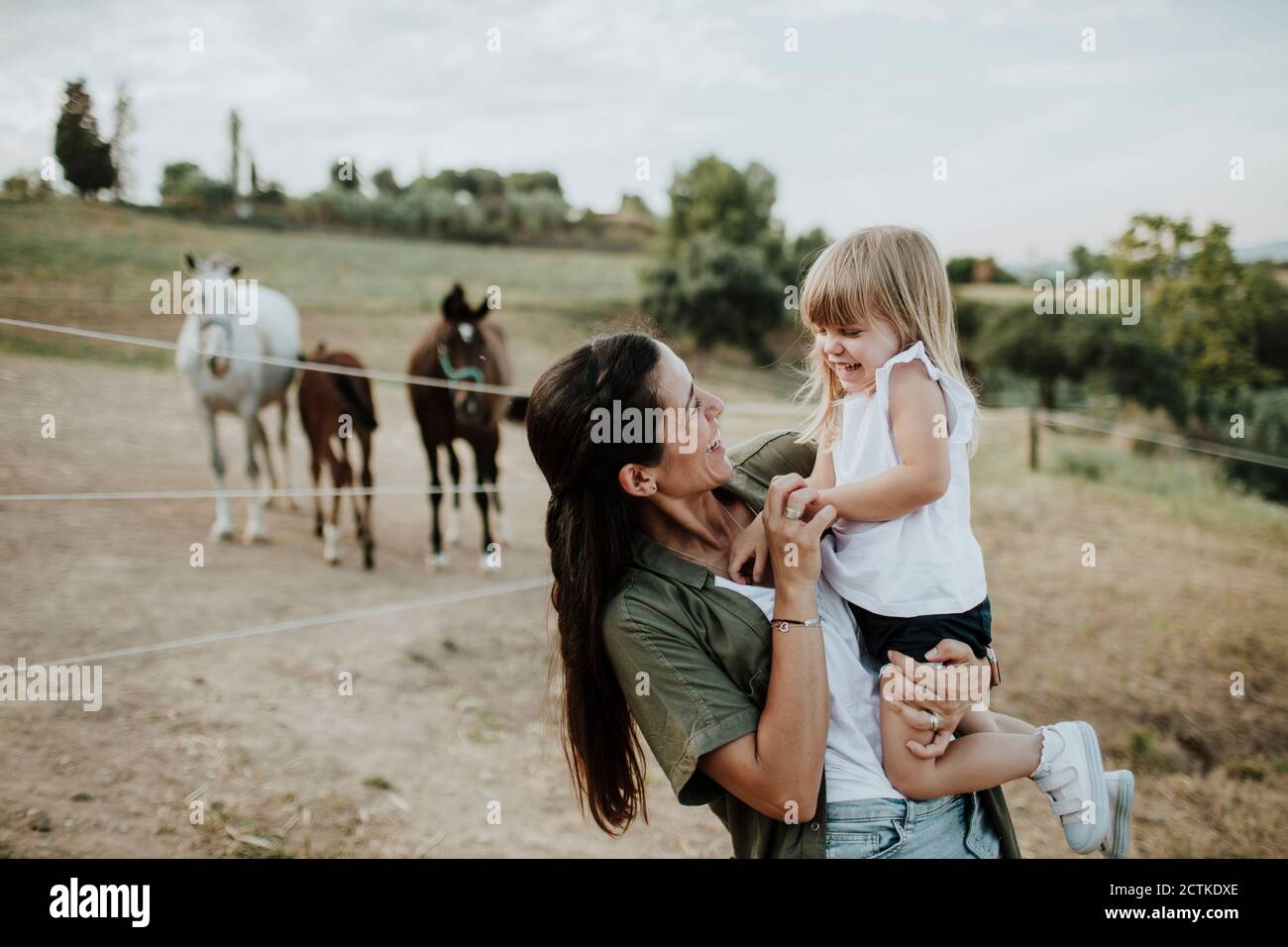 Madre sorridente che porta la figlia allegra mentre si trova contro i cavalli terra Foto Stock