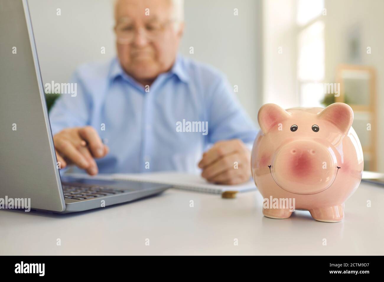 Banca piggy in piedi sulla scrivania con l'uomo anziano offuscato che usa portatile per pagare le fatture online in background Foto Stock