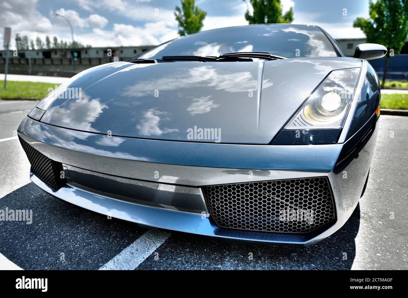 Vista frontale isolata e grandangolare di una potente auto sportiva di colore metallizzato blu parcheggiata su una strada pubblica. Il cielo e le nuvole si riflettono sul metallo lucido Foto Stock