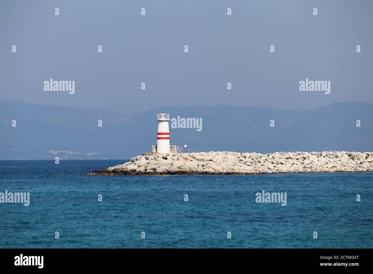 Faro torre su un molo di pietra in mare azzurro contro montagne misty Foto Stock