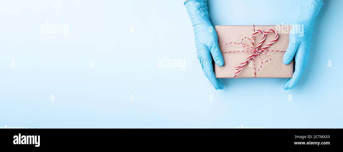 Mani in guanti medici che tengono scatola regalo di Natale su sfondo blu. Concetto di sicurezza gifting. Foto Stock