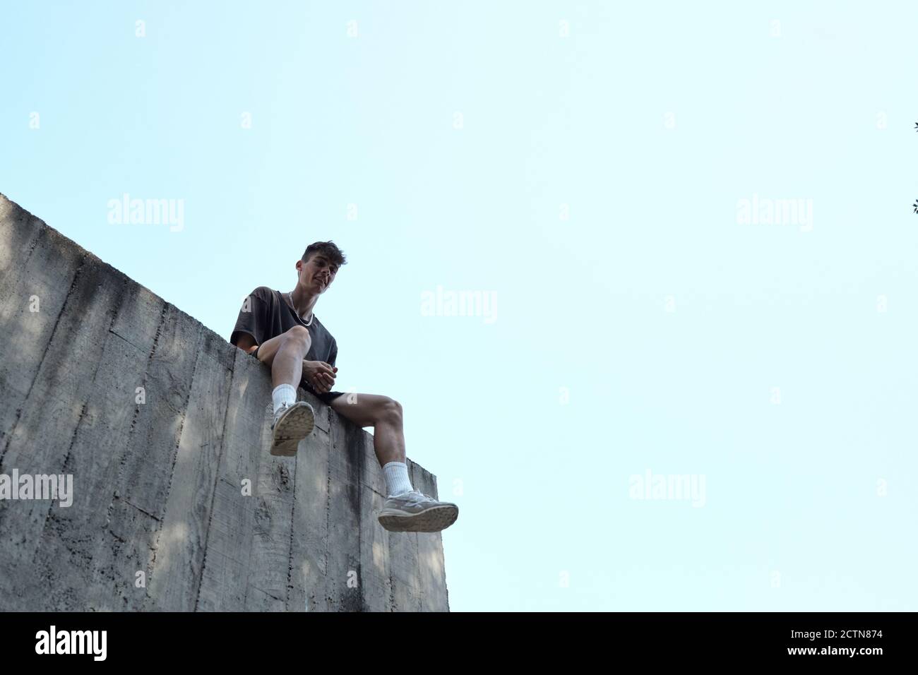 Da sotto di sorridente maschio seduto su parete alta di edificio in cemento e relax durante l'allenamento al parco in città Foto Stock