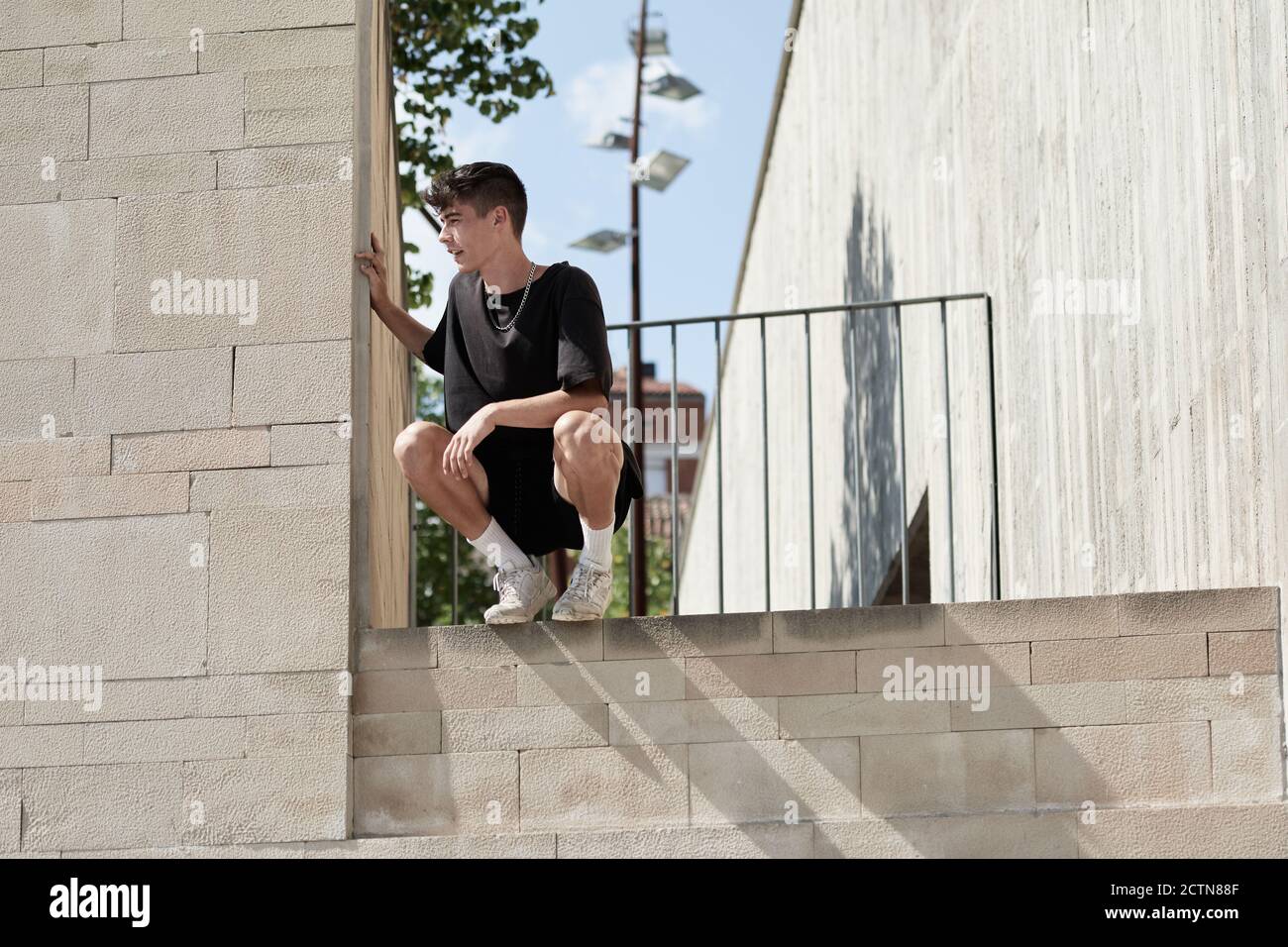 Da sotto di sorridente maschio seduto su parete alta di edificio in cemento e relax durante l'allenamento al parco in città Foto Stock