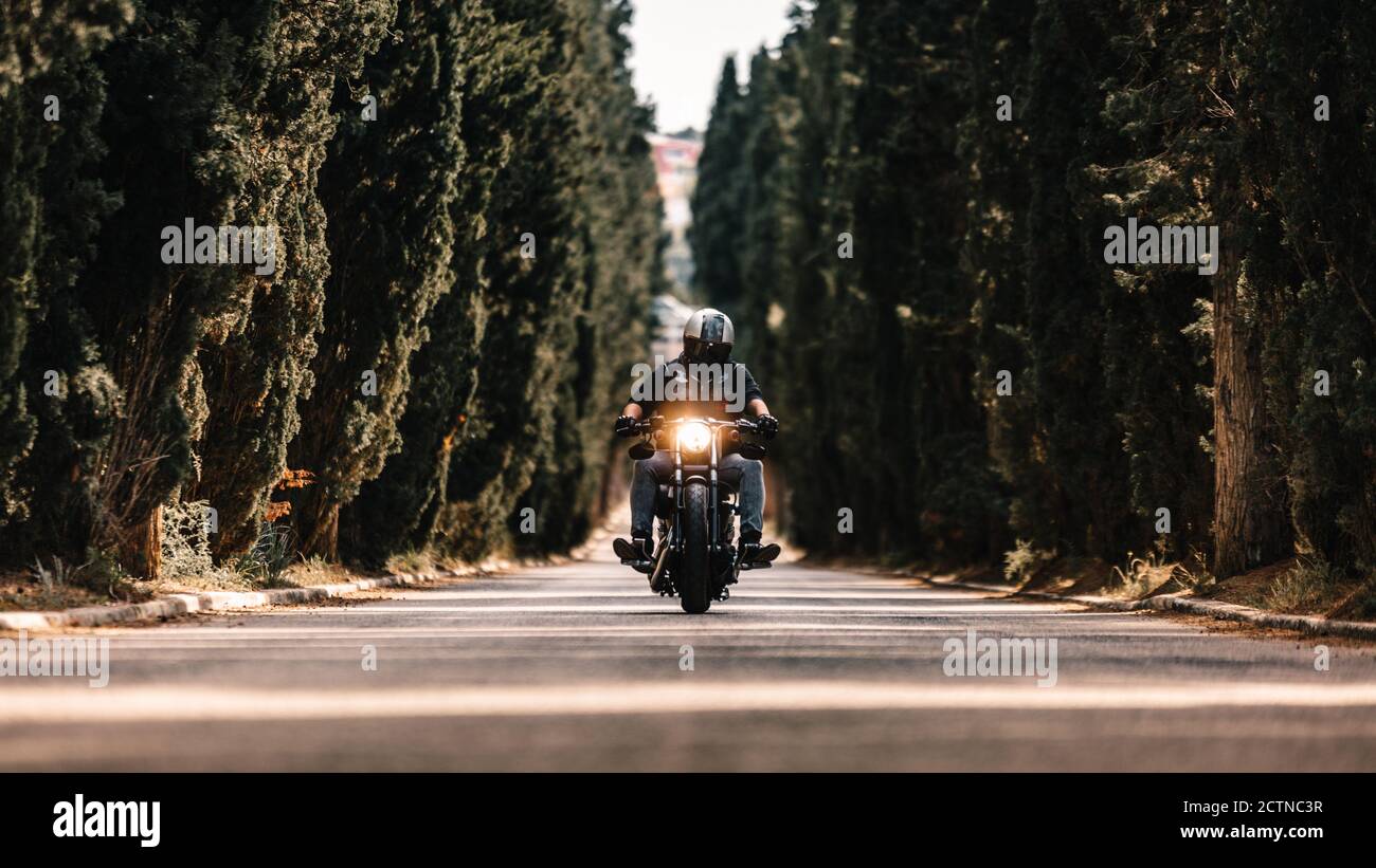 Motociclista in giacche di pelle nera e casco che cavalcano moto potente su strada asfaltata che conduce tra verde foresta in campagna Foto Stock