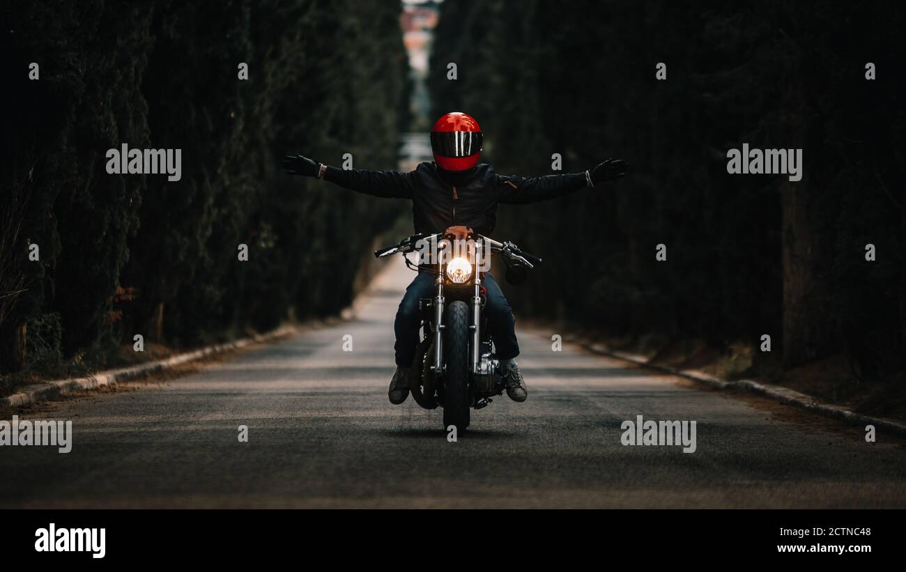 Biker con giacche in pelle nera e casco a braccia aperte guida potente moto su strada asfaltata che conduce tra foresta verde in campagna Foto Stock