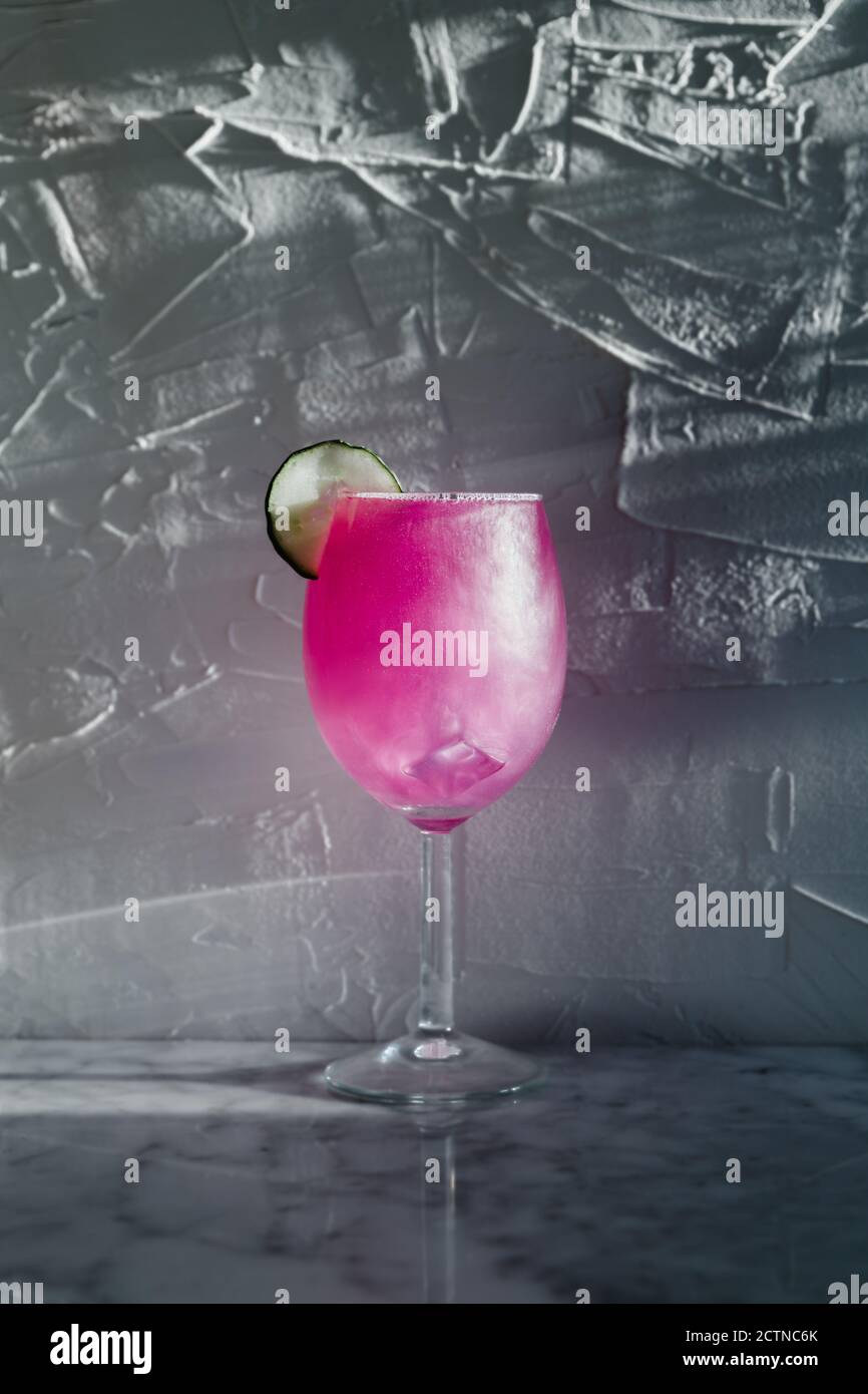 Wineglass di rinfrescante cocktail viola chiaro guarnito con erbe e. lime affettato posto alla luce del sole contro sfondo grigio offuscato Foto Stock