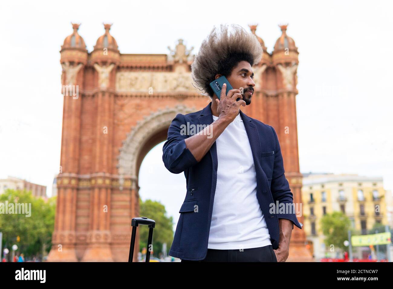 Giovane uomo nero in tuta e valigia in piedi per strada parlando con il suo cellulare Foto Stock