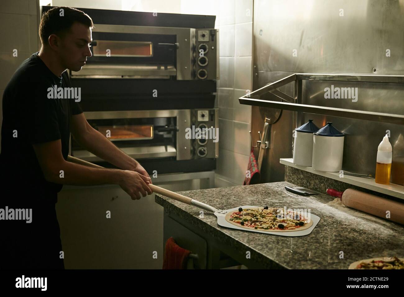 Vista laterale dello chef maschile con pala in metallo che prende  tradizionale Pizza crude italiana preparata per la cottura dal piano in  marmo un forno per la cottura in cucina del ristorante