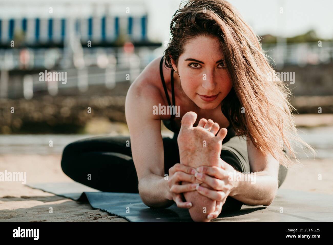 Sorridente donna flessibile seduta sul tappetino a Janu Sirsasana e. fare la curva in avanti mentre si guarda la macchina fotografica e si pratica yoga in spiaggia in estate Foto Stock