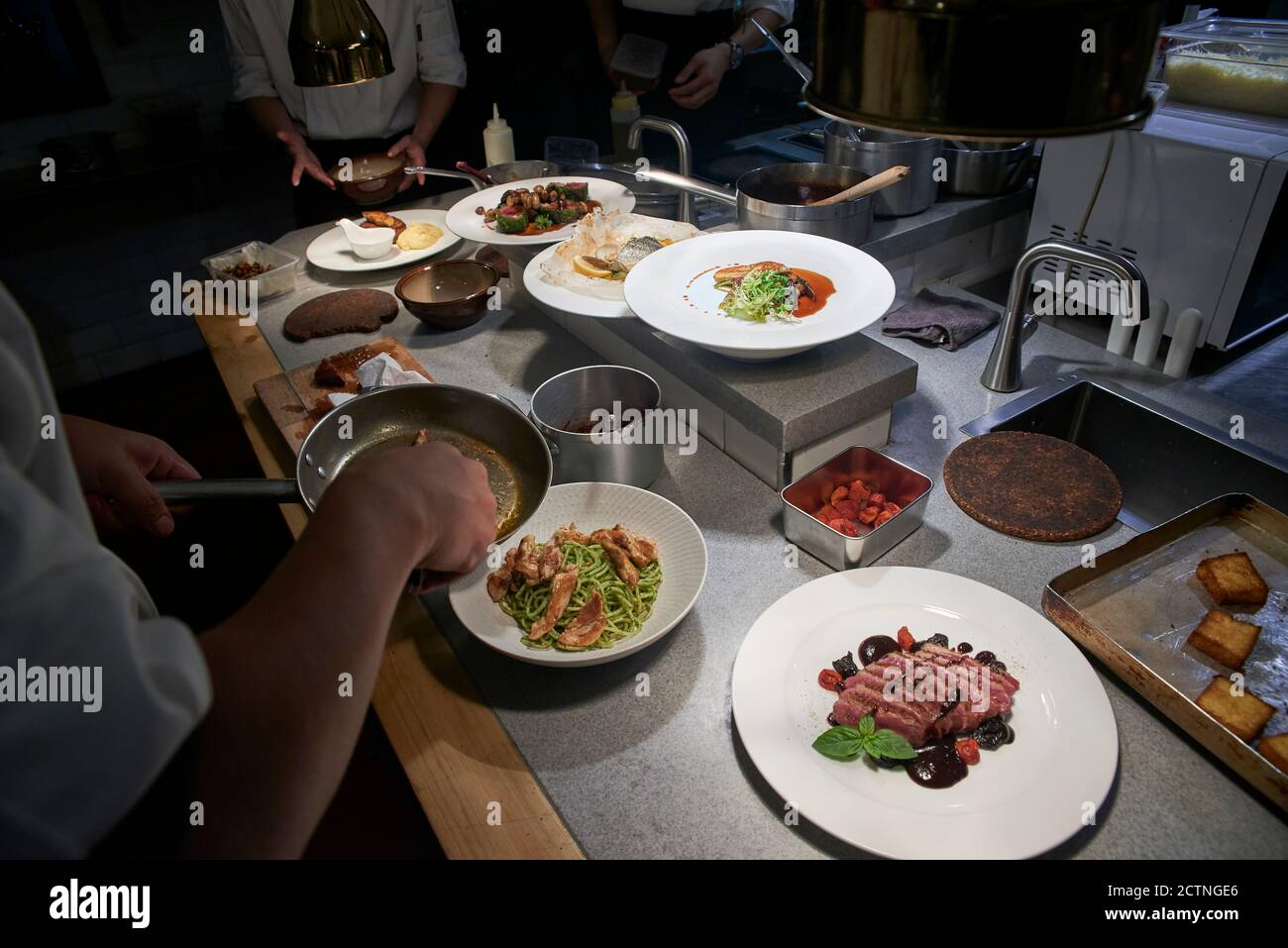 Alto angolo di raccolto anonimo chef aggiungendo filetti di pollo fritti nel piatto con la pasta mentre prepara il cibo con i colleghi dentro cucina moderna del ristorante Foto Stock