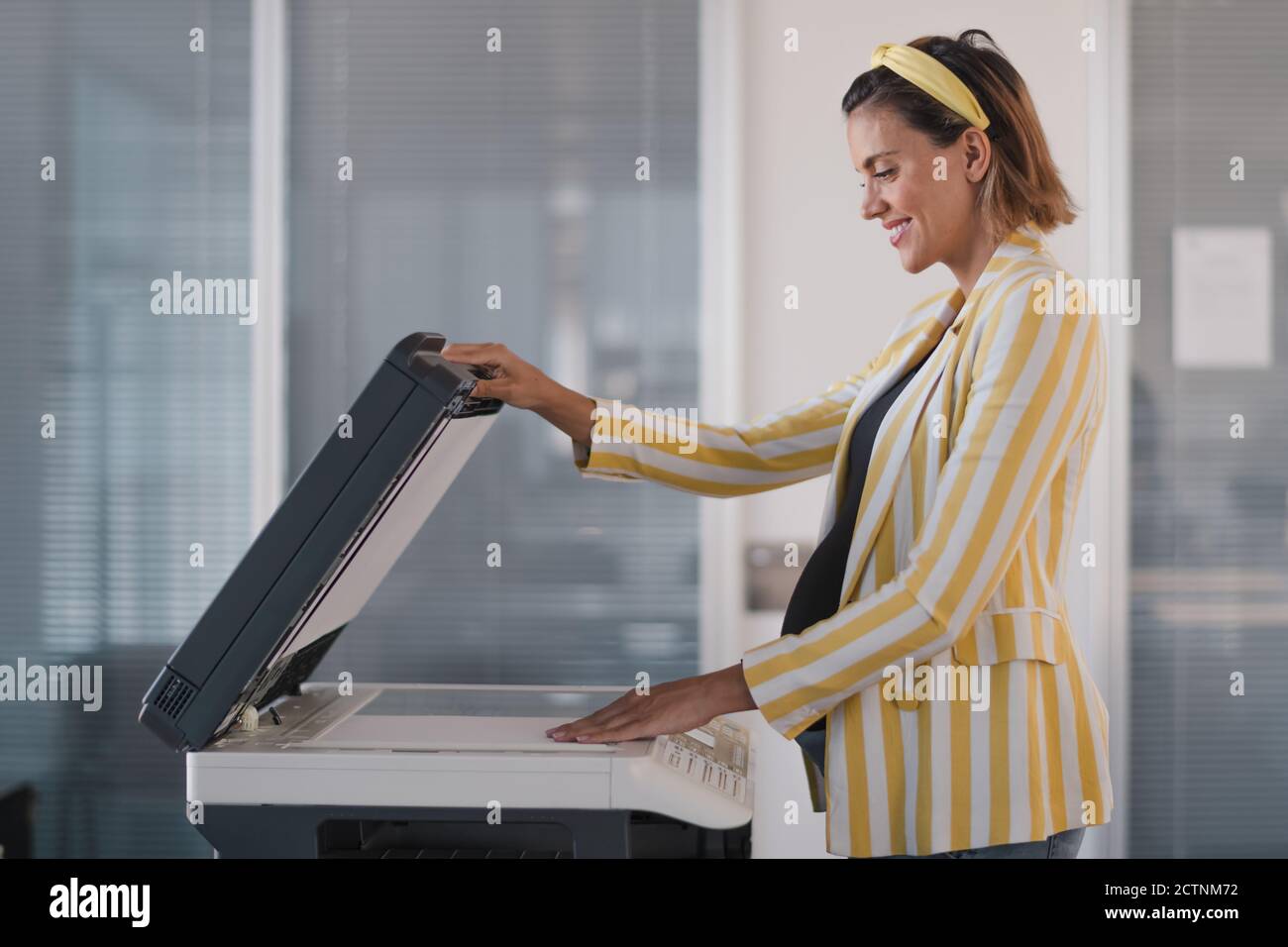 Vista laterale di una donna d'affari incinta ottimista in elegante vestito mettendo foglio di carta su fotocopiatrice mentre lavora in uno spazio di lavoro moderno Foto Stock