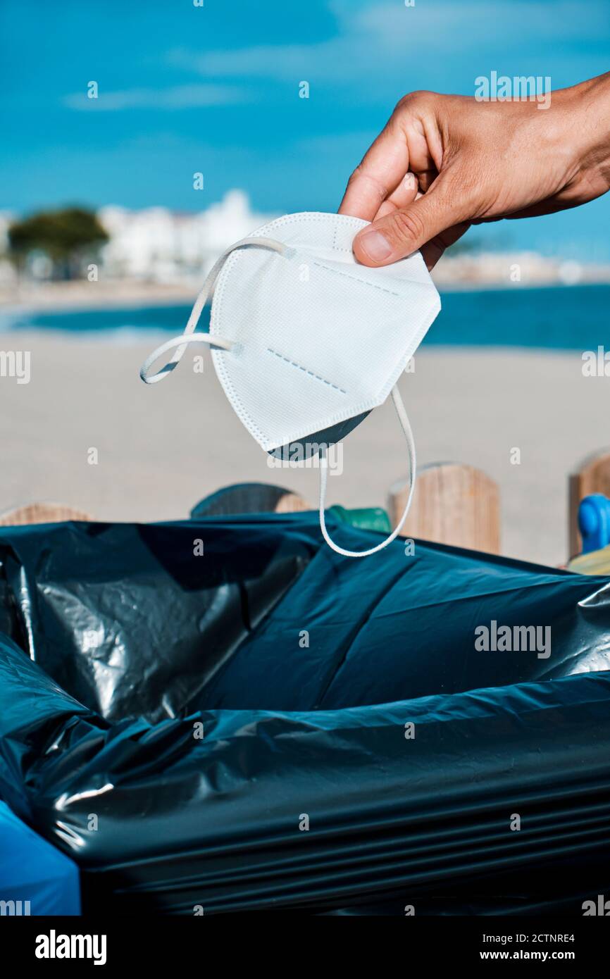 closeup di un uomo caucasico che lancia un usato bianco monouso maschera al cestino vicino alla spiaggia Foto Stock