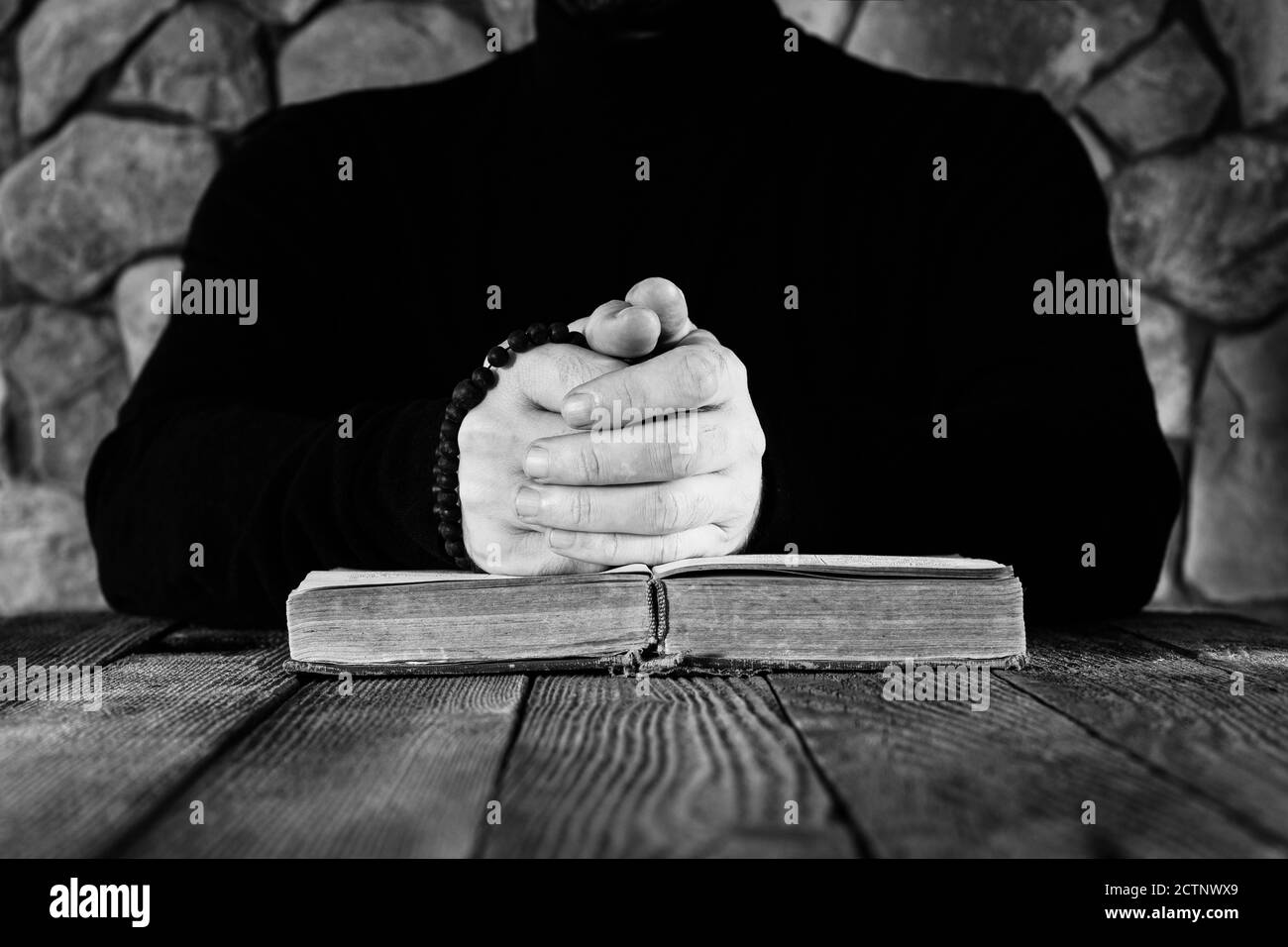 Un uomo in abiti neri con un rosario in mano davanti ad un vecchio libro aperto la Bibbia o il Corano, il concetto di preghiera, foto in bianco e nero Foto Stock