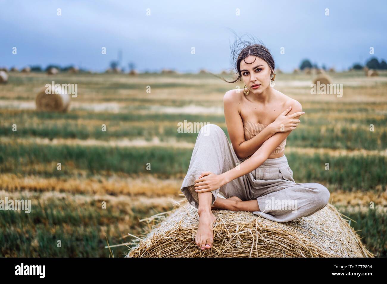 Bruna in mutande di lino e spalle nude seduta su balle di fieno in caldo  giorno d'autunno. Donna che guarda la macchina fotografica. Dietro di lei  c'è un campo di grano Foto