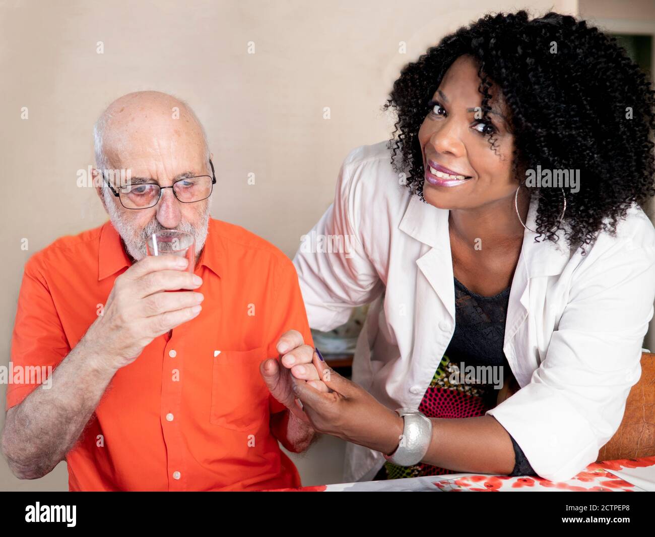 Ritratto orizzontale di un'infermiera afroamericana che tiene le mani con un uomo edero e facendolo bere un bicchiere di acqua Foto Stock