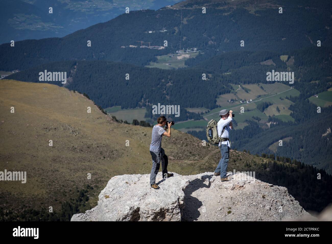 Due uomini si trovano su uno sperone roccioso per fotografare la vista delle Dolomiti sopra la Val Gardena. Foto Stock