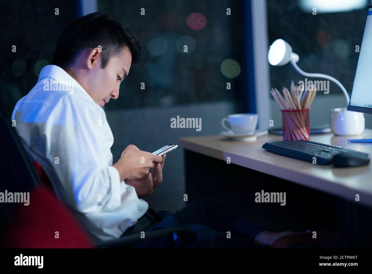 Lavoratore in ufficio, giovane uomo tailandese che guarda lo smartphone in ufficio a tarda notte. Concetto di lavoro notturno. Foto Stock