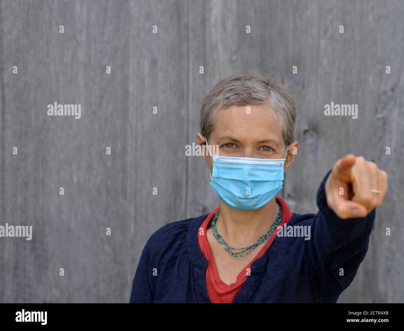 La donna caucasica di mezza età indossa una maschera chirurgica blu e punta con il dito indice verso lo spettatore durante la pandemia della corona del 2019/20. Foto Stock