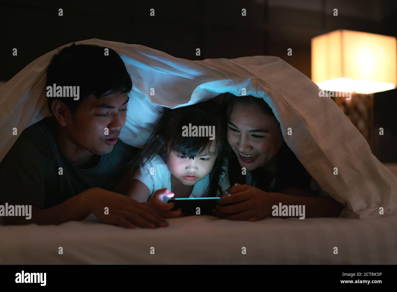 Asian Happy famiglia madre, padre e piccola figlia guardando film o cartoni animati in smartphone insieme e coperta coprire la testa a letto di notte a. Foto Stock