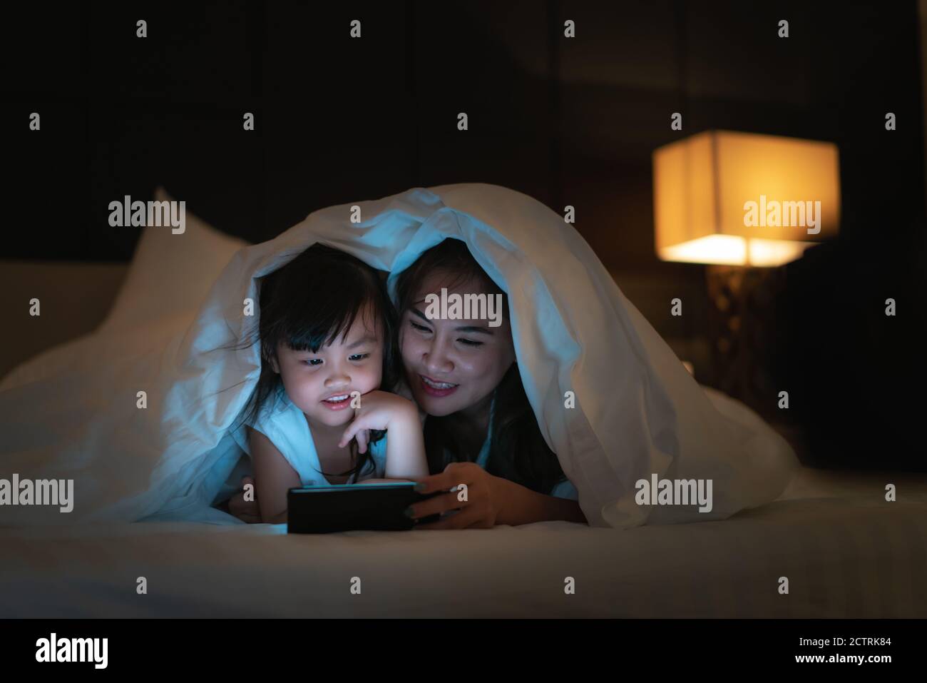 Asian felice madre di famiglia e piccola figlia che guarda il film o. cartone animato in smartphone insieme e coperta coprire la loro testa in letto di notte a casa Foto Stock