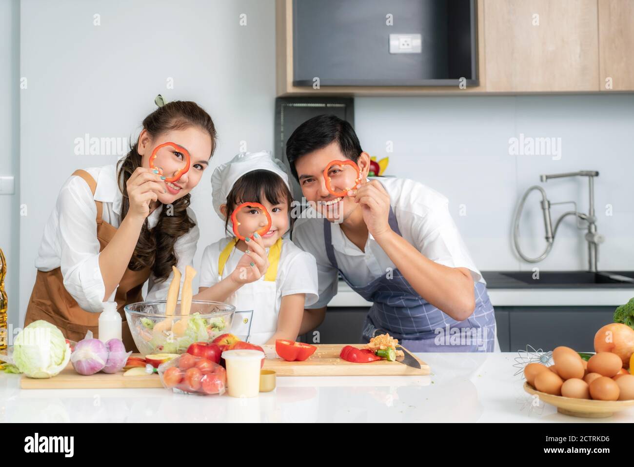 La famiglia asiatica con il padre, la madre e la figlia carina della bambina nei cappelli dello chef stanno divertendosi mentre cucinando in cucina a casa Foto Stock