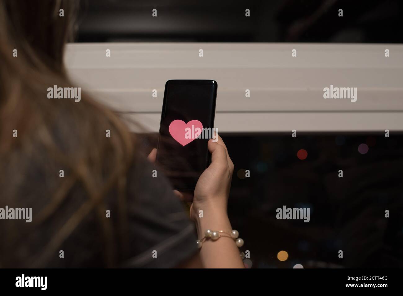 Donna di fronte a una finestra che tiene il telefono cellulare con un cuore sullo schermo Foto Stock