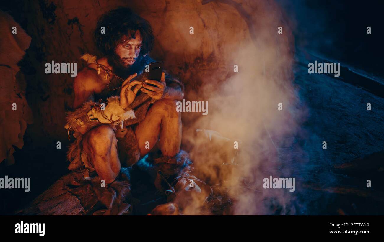 Tribù di Preistic, cacciatore primitivo Gatherer che indossa pelle animale utilizza smartphone in una grotta di notte. Neanderthal Homo Sapiens Male Browsing Foto Stock