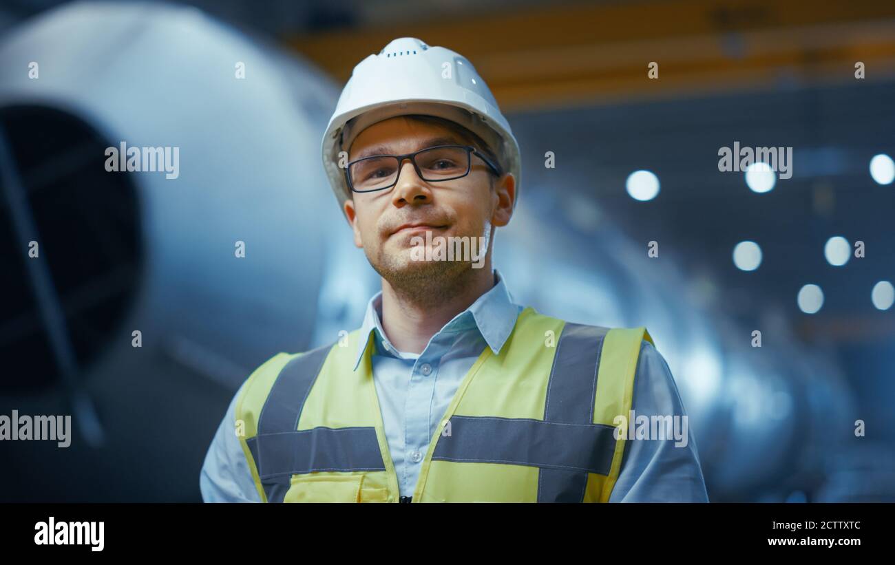Ritratto di giovane professionista ingegnere del settore pesante lavoratore indossando giubbotto di sicurezza e Hardhat sorridendo sulla fotocamera. In background non messo a fuoco grande Foto Stock