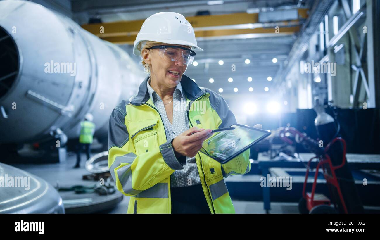 Professionista Heavy Industry femmina Engineer indossare l'uniforme di sicurezza, tiene il Tablet computer digitale e spiega la progettazione del prodotto. Fabbrica industriale Foto Stock