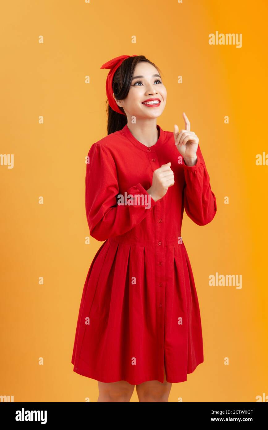 Sorpresa bella giovane donna in vestito rosso sta indicando. Studio di tre quarti di lunghezza su sfondo arancione. Foto Stock