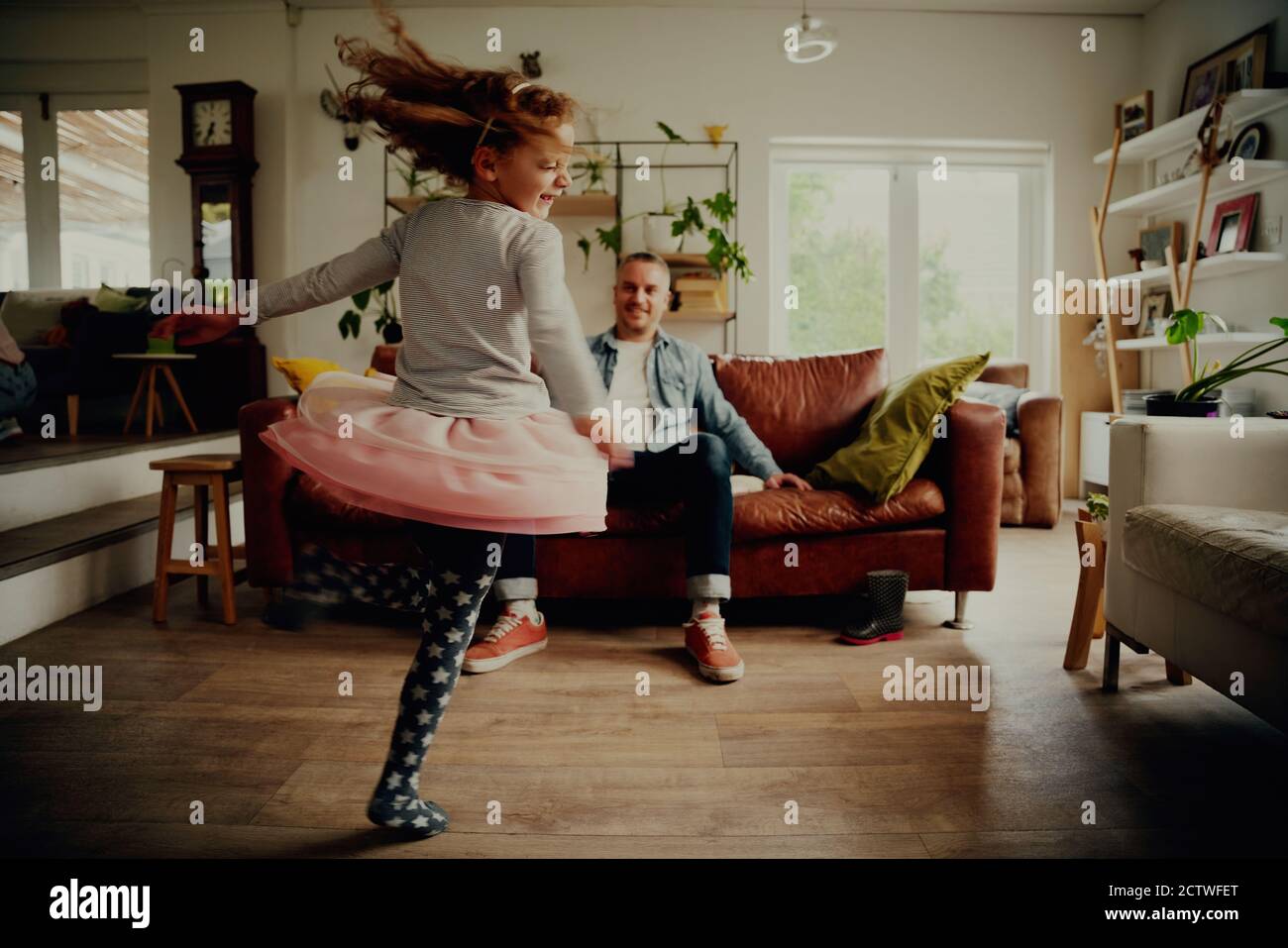 Ragazza attiva che danzano e roteano in soggiorno con padre guardare mentre si rilassa sul divano a casa Foto Stock