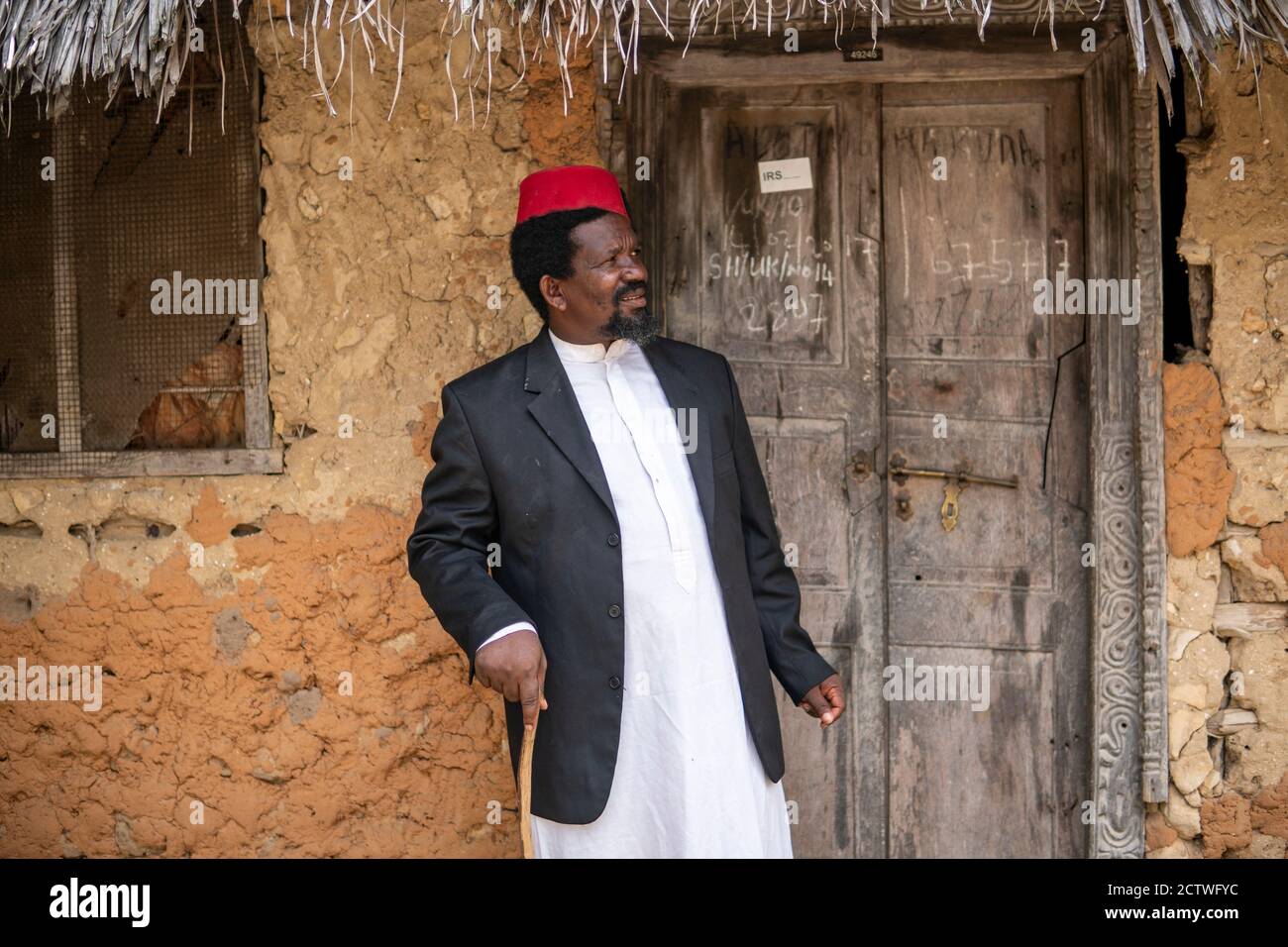 Un uomo anziano africano nel cappello rosso musulmano Taqiyyah Fez E Blazer  su vestito bianco muovendo un bastone per zoppo Persone vicino al Rifugio  base con tetto in paglia Foto stock -