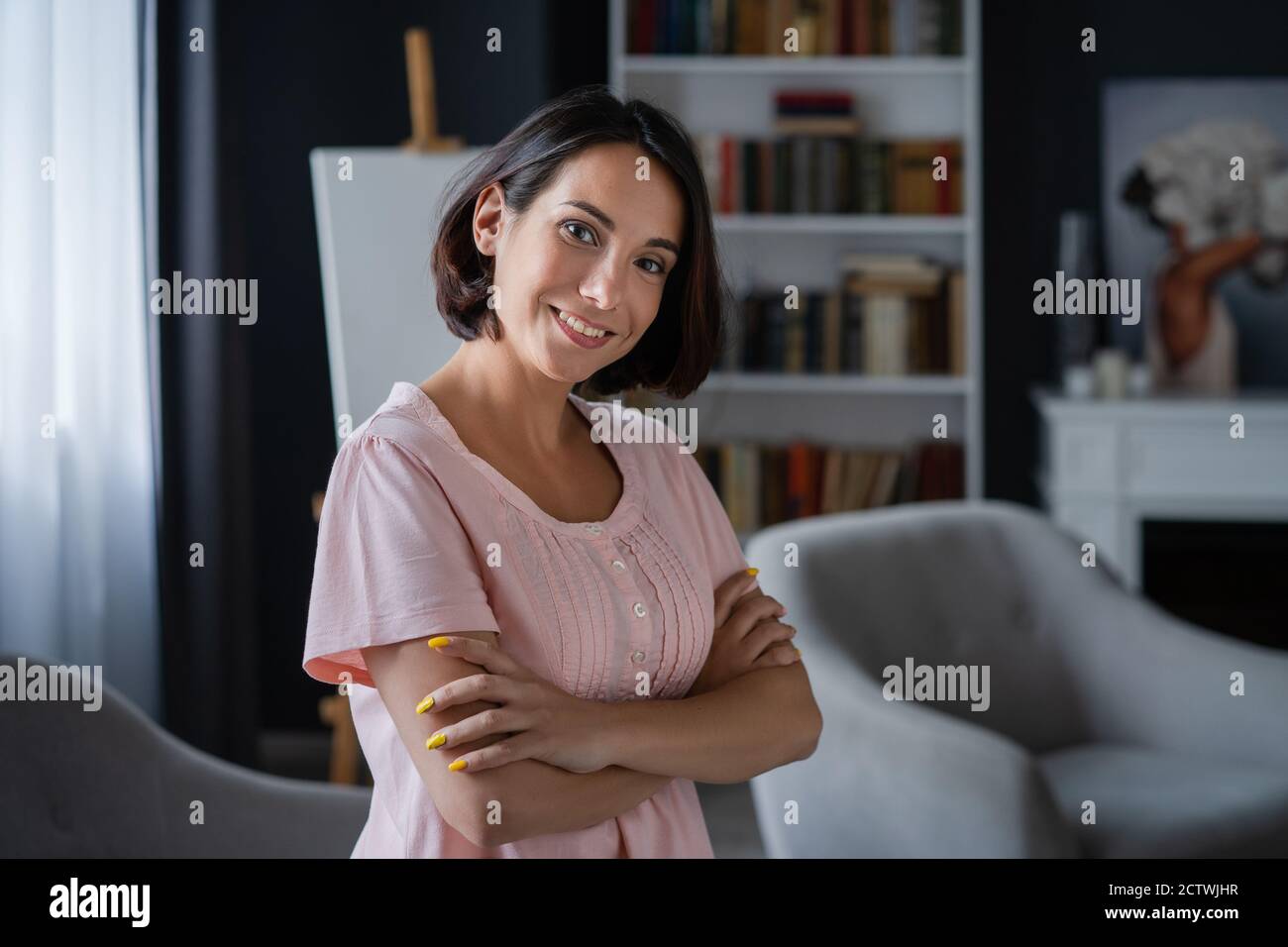 La giovane donna si alza nella stanza con le braccia ripiegate sul petto. Sullo sfondo c'è un cavalletto, una libreria e una sedia. Foto di alta qualità Foto Stock
