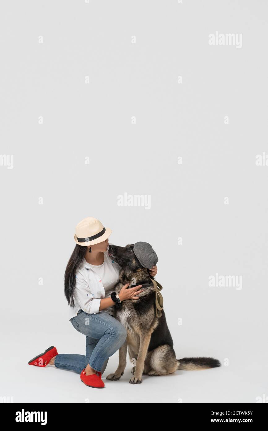 Donna in cappello bacia il cane da pastore in cappuccio contro uno sfondo isolato. Il proprietario si siede con il cane in studio. Foto di alta qualità Foto Stock