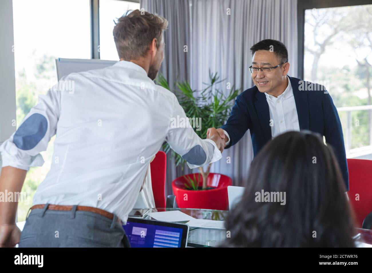 Uomo d'affari asiatico e caucasico in piedi scuotendo le mani sul tavolo in una riunione di affari, con i colleghi seduti accanto a loro. Business creativo Foto Stock
