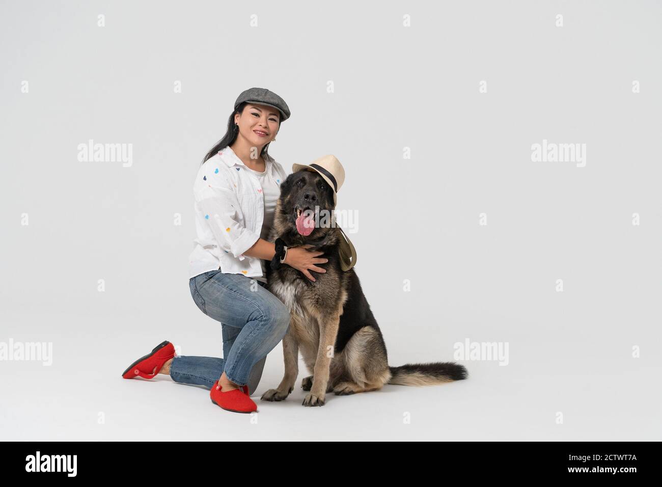 Donna asiatica in un cappello con un Pastore dell'Europa orientale in un cappello si siede sul pavimento dello studio. Proprietario e cane in cappelli su uno sfondo isolato. Alta Foto Stock