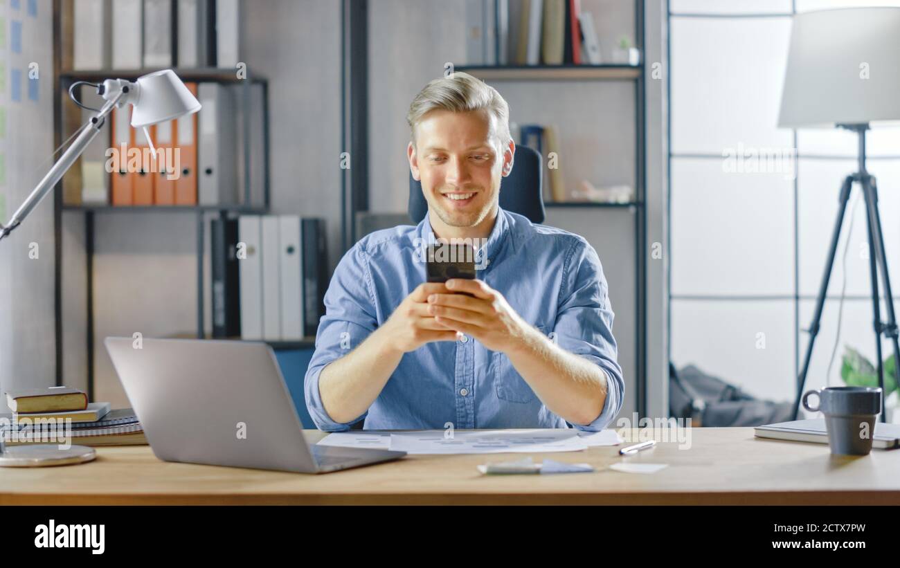 Imprenditore creativo seduto alla sua scrivania funziona su un laptop in un elegante ufficio, utilizza smartphone per le applicazioni di social media, e-mail Business Associates Foto Stock