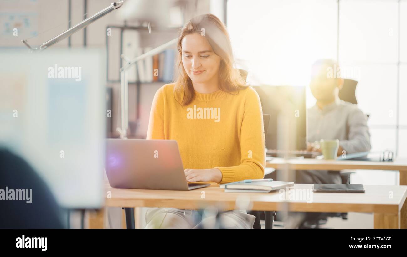 Bella e creativa giovane donna seduta alla sua scrivania utilizzando un computer portatile. In background Bright Ufficio dove diversi team di giovani Foto Stock