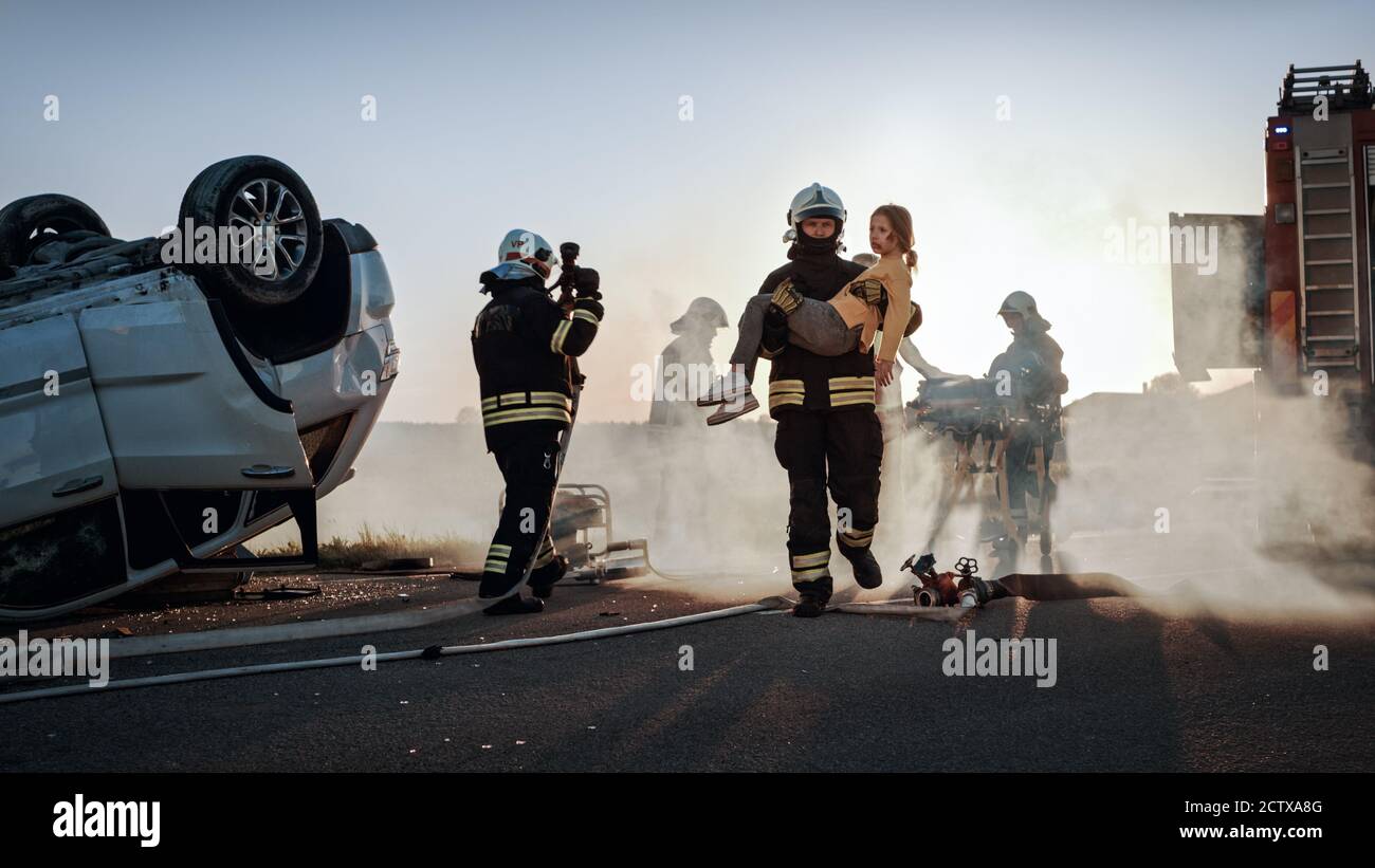 Brave Firefighter trasporta la giovane ragazza ferita alla sicurezza dove si riunì con la sua madre amorevole. In background Auto Crash incidente stradale Foto Stock