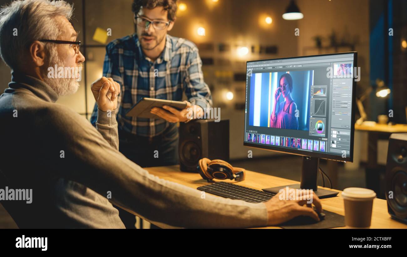 In serata Creative Middle Aged Photo Editor lavora su un computer desktop utilizza il software di editing delle immagini, ha conversazione con uno Specialista più giovane che Foto Stock
