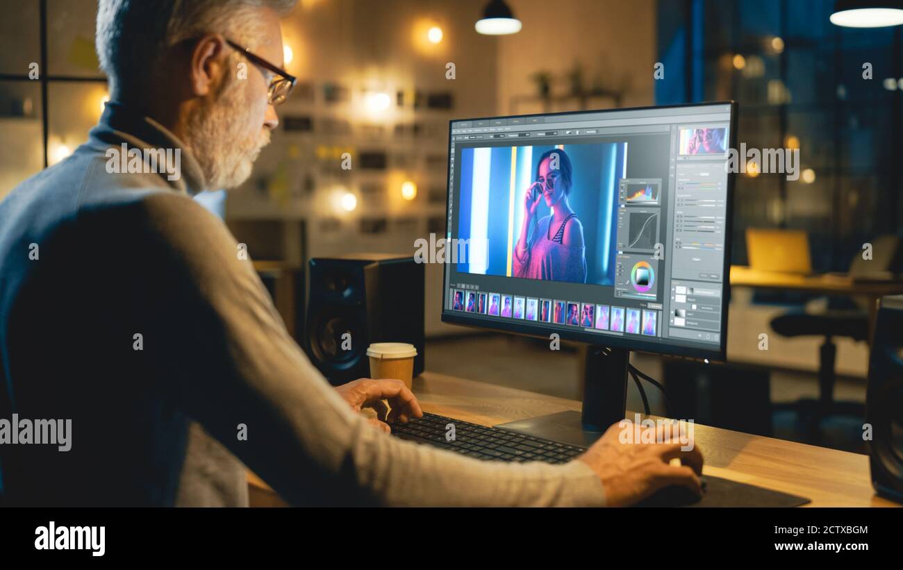 In tarda serata Creative Middle Aged Photo Editor funziona su un computer desktop utilizza il software di editing delle immagini. Ufficio moderno ed elegante in serata Foto Stock