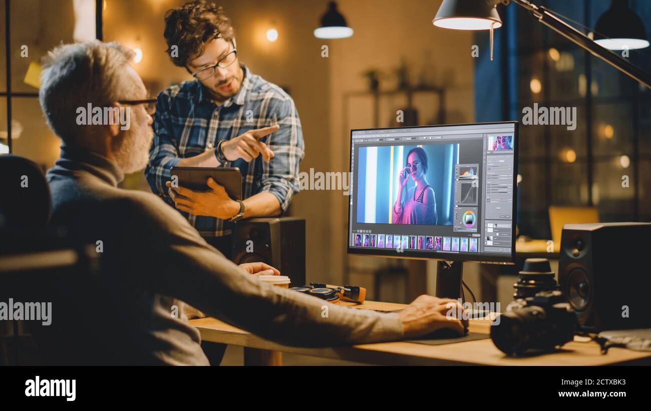 In serata Creative Middle Aged Photo Editor lavora su un computer desktop utilizza il software di editing delle immagini, ha conversazione con uno Specialista più giovane che Foto Stock