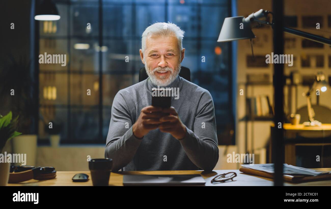 Ritratto del bell'uomo d'affari di media età Bearded usa lo smartphone mentre si siede alla sua scrivania, ride e sorride. Lavoro da Foto Stock