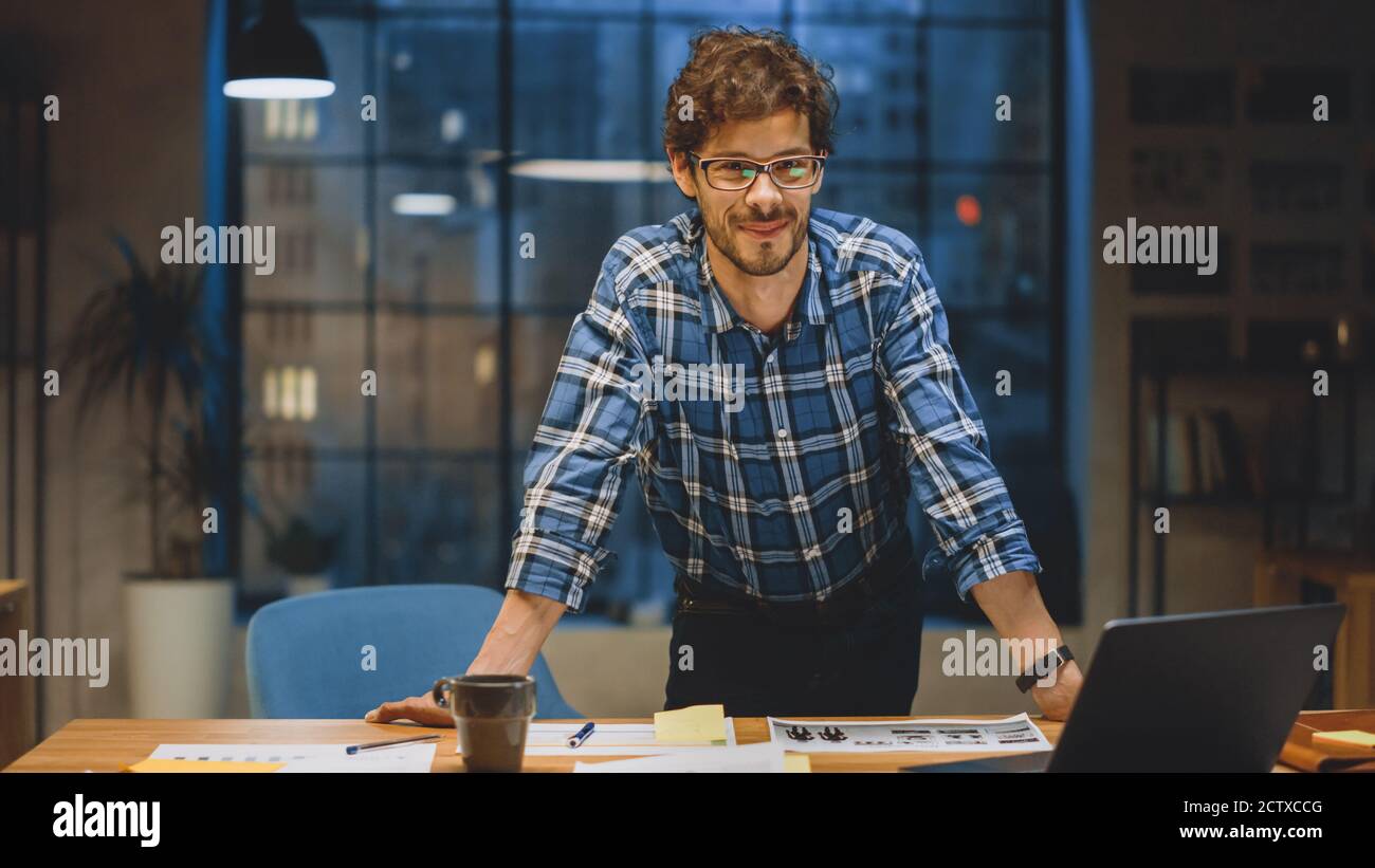 Young Creative Designer che lavora alla sua scrivania, sorride su Camera. Sta facendo la documentazione, scegliendo il disegno, il concetto e gli schizzi giusti. In background Foto Stock