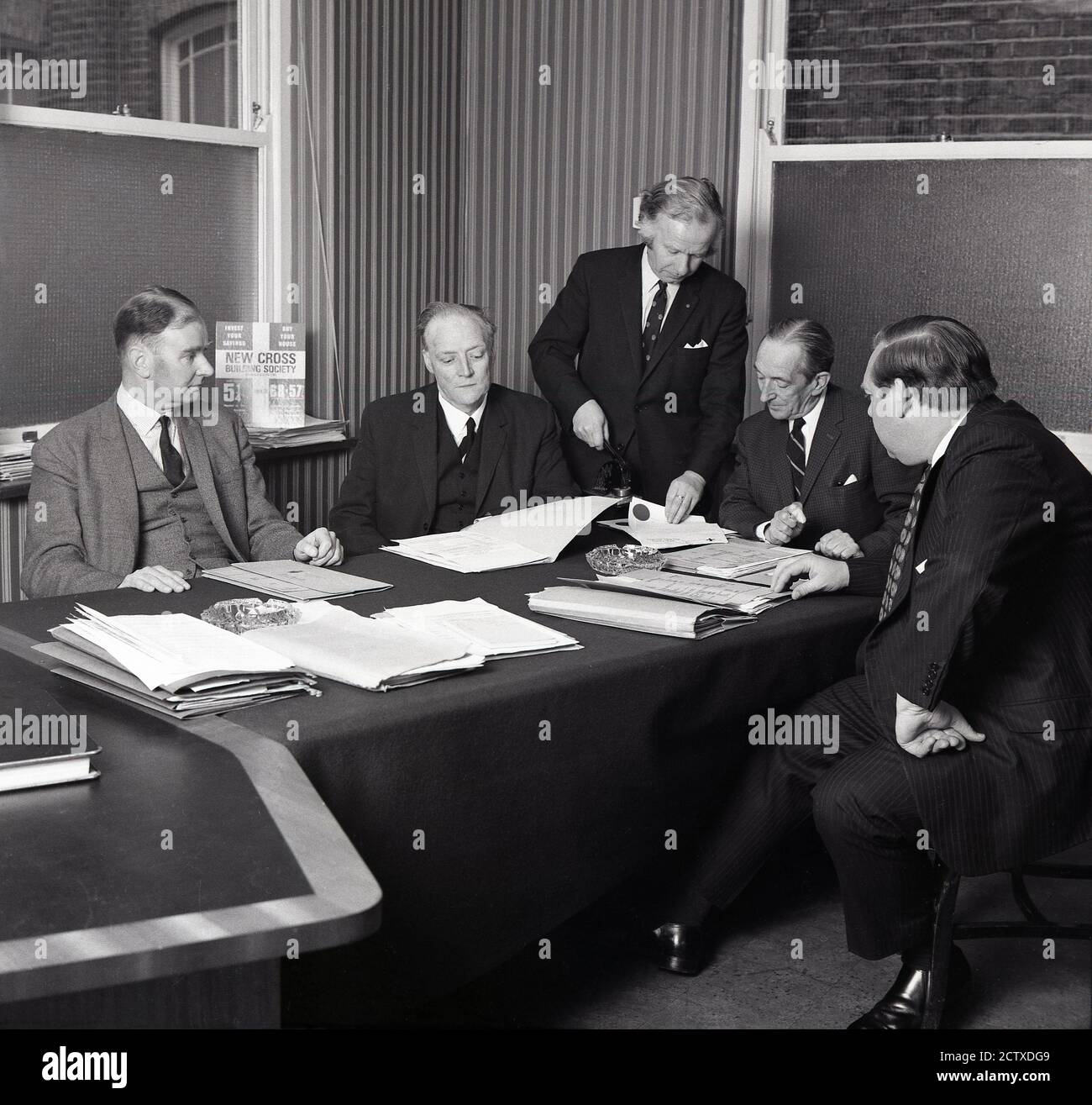 Anni '1960, storico, in una sala riunioni, i dirigenti senior del settore bancario si riuniscono intorno a un tavolo coperto in feltro, mentre uno di loro mette un sigillo ufficiale su alcuni documenti, South London, England, UK. Foto Stock