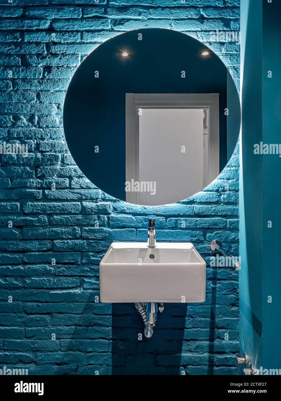 Bagno con parete in mattoni blu, lavabo quadrato e specchio rotondo. Interni minimalisti. Foto Stock