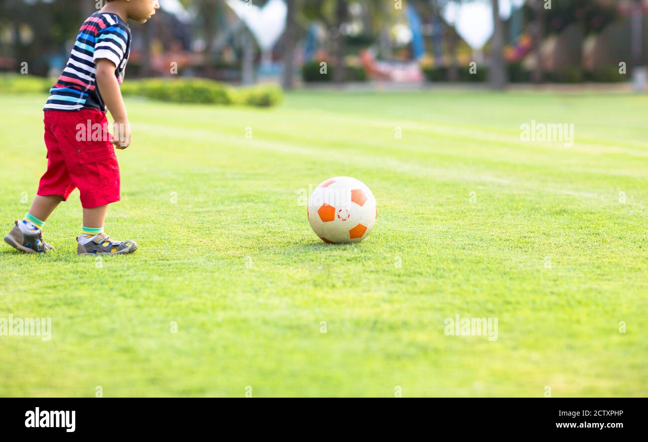 Il ragazzino imparando a giocare a calcio preparandosi a calciare la palla sull'erba in un parco in una giornata estiva. Foto Stock
