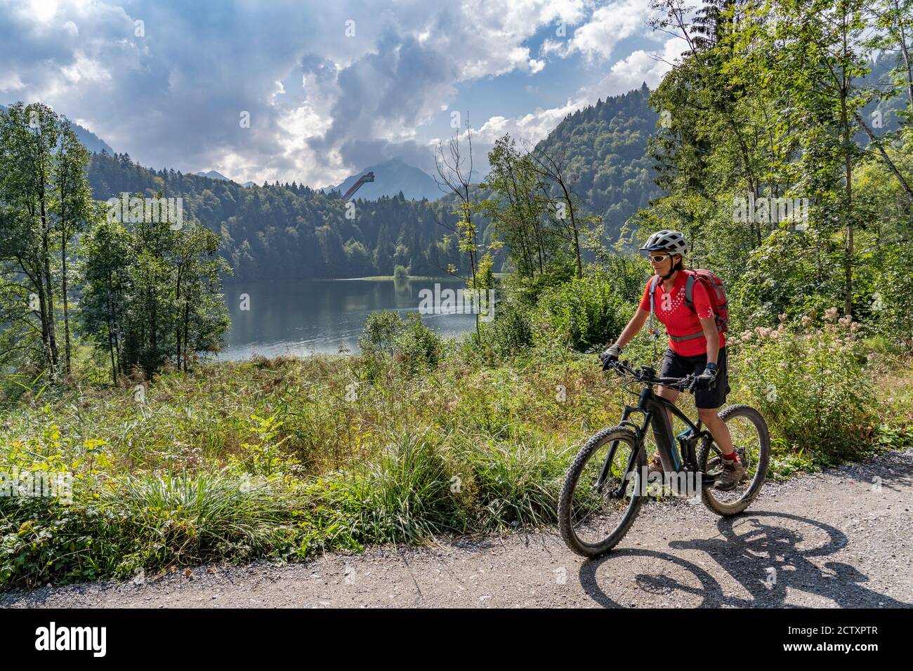 Bella donna anziana che guida la sua mountain bike elettrica sopra il lago Freiberg e una grande collina di volo sci nelle Alpi di Allgau vicino Oberstdorf, Germania Foto Stock