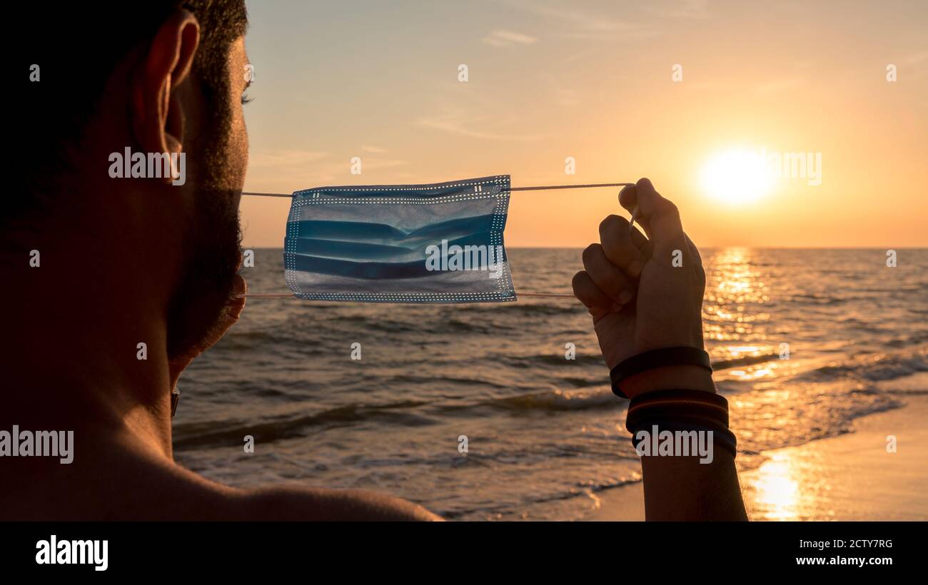 Uomo spagnolo caucasico in maschera medica durante il blocco coronavirus a bella spiaggia con luce tramonto. Ritratto post covid. Persone in sicurezza Foto Stock