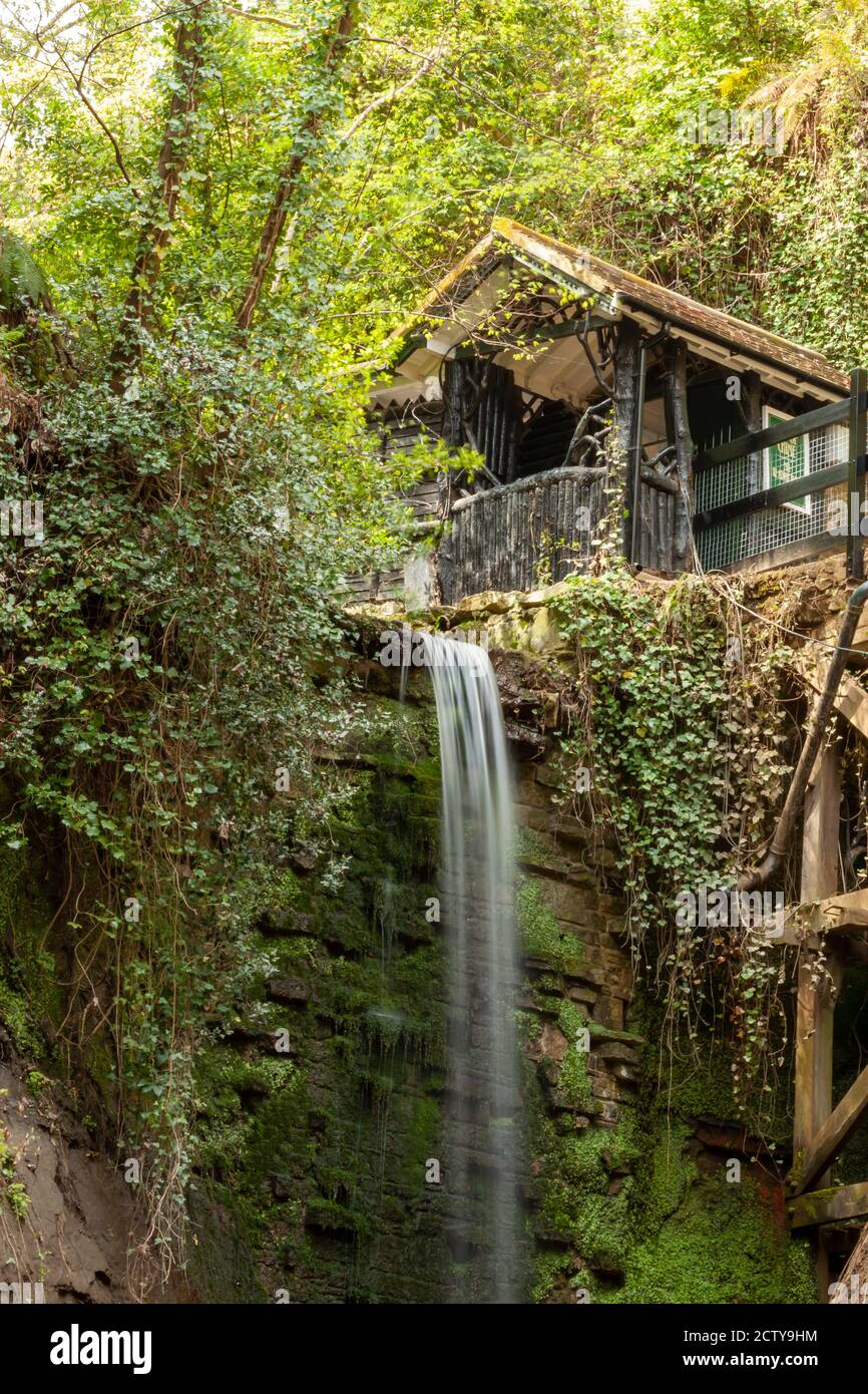Immagine a lunga esposizione di una piccola cascata. C'è una casa di legno sulla cima della scogliera al bordo della parete rocciosa. Il muro di pietra dietro la caduta è Foto Stock